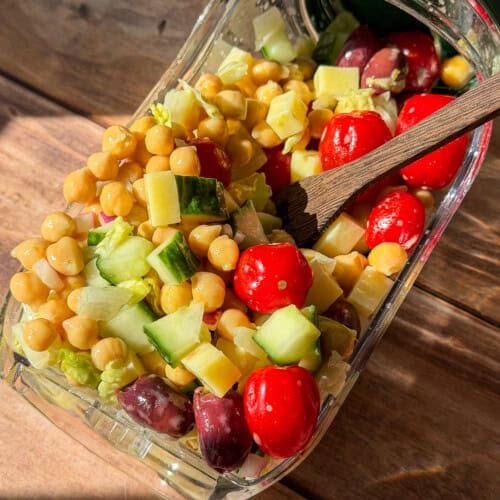 A glass container tipped on its side spills a colorful salad with chickpeas, grape tomatoes, cucumber, cheese cubes, and olives onto a wooden surface. A wooden fork rests inside the container.
