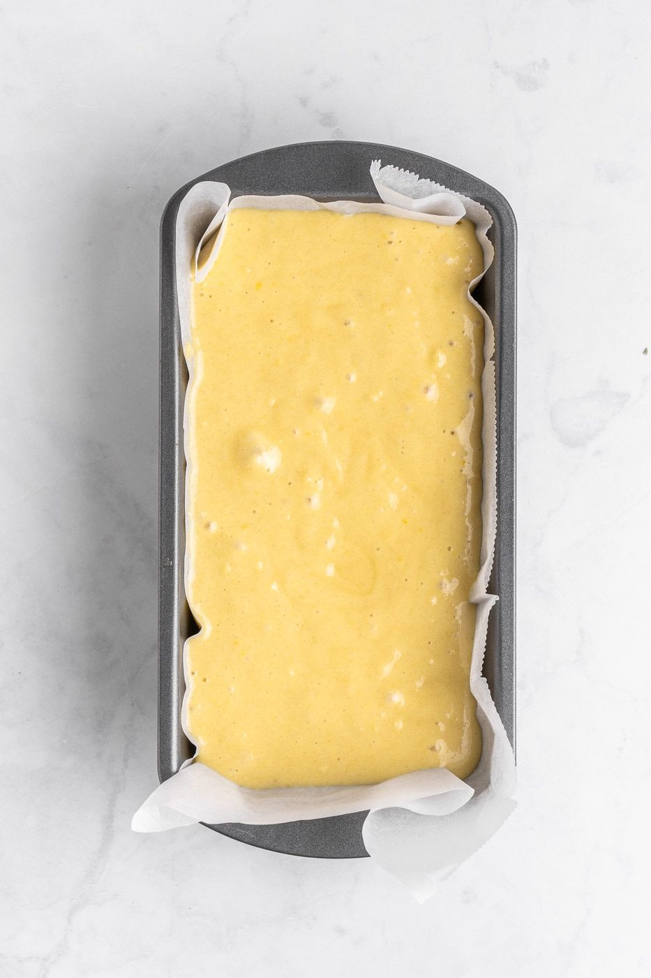 A loaf pan lined with parchment paper is filled with uncooked yellow batter, ready to be baked, set against a white marble background.