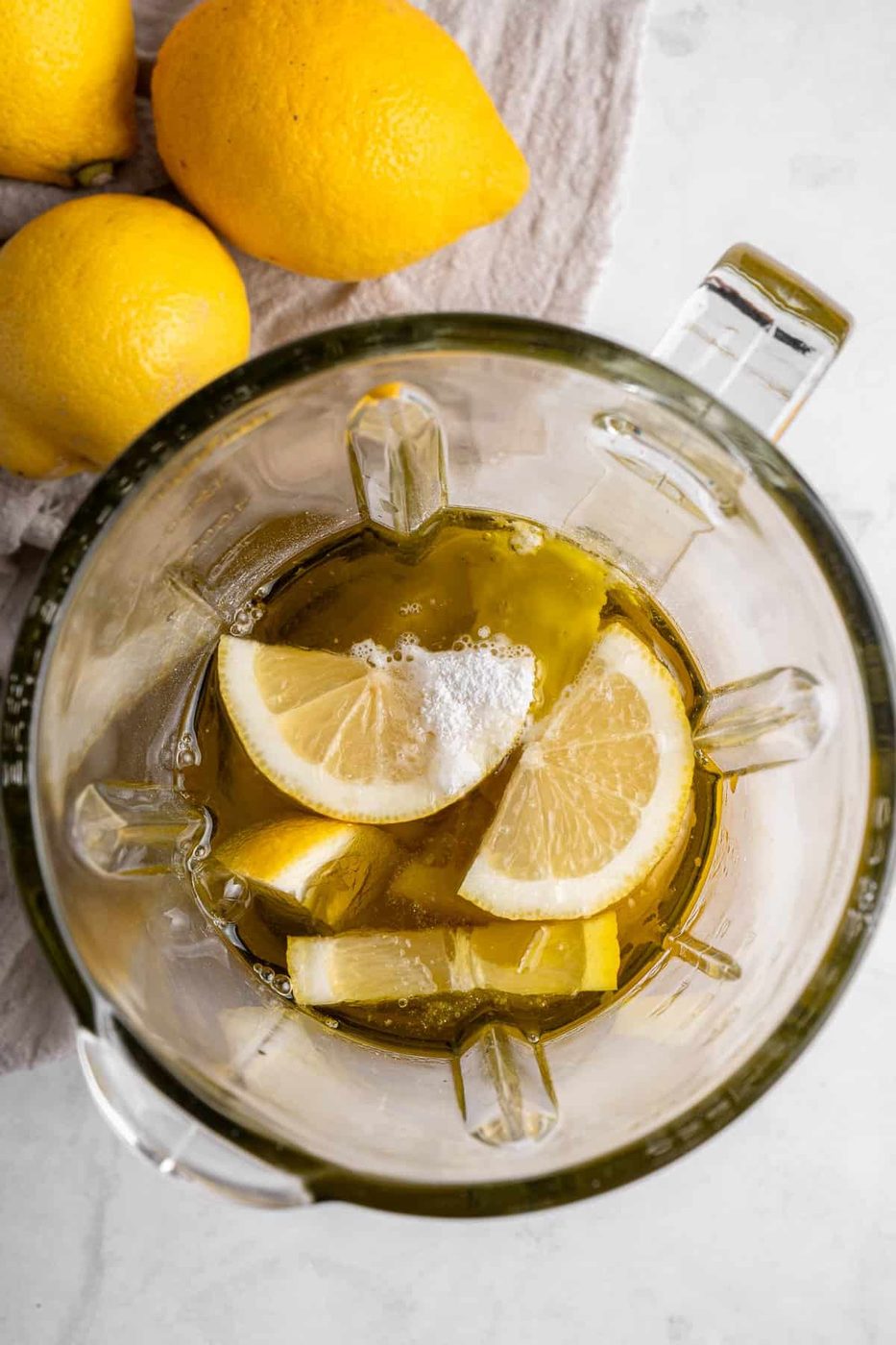 Top view of a blender containing lemon slices, olive oil, and a sprinkle of salt, placed on a light-colored surface. Three whole lemons rest beside the blender on a textured cloth.