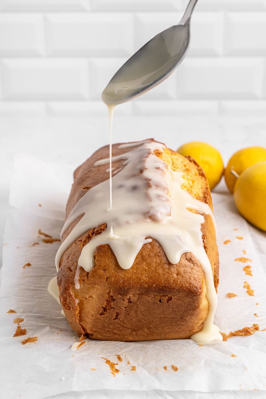 A loaf of lemon cake on parchment paper is being drizzled with icing from a spoon. Two whole lemons are placed in the background. The setting is a white kitchen counter with a tiled backsplash.