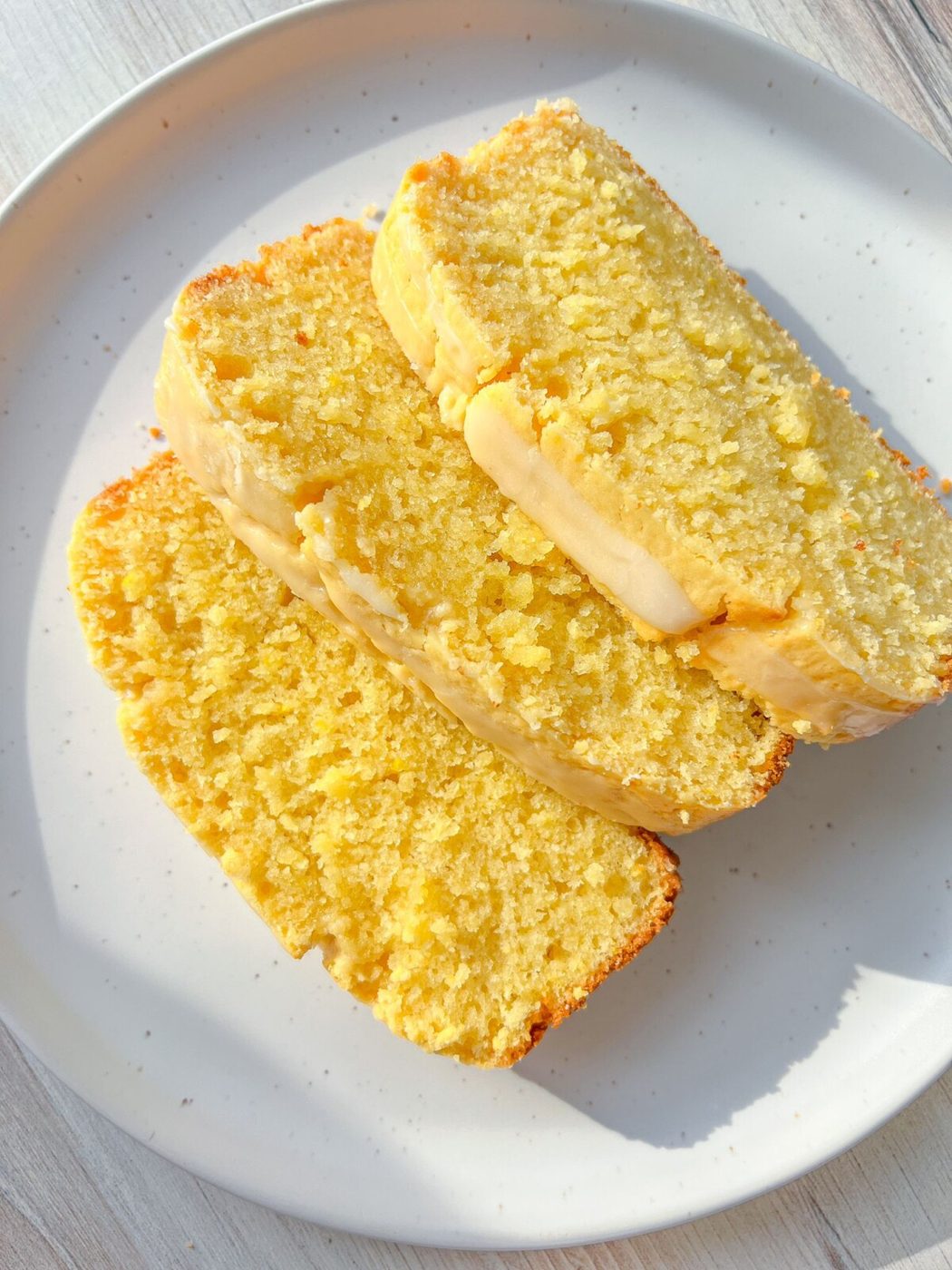 Three slices of moist Whole Lemon Blender Cake arranged on a white plate, with a light glaze visible on the surface, set on a light-colored tabletop.
