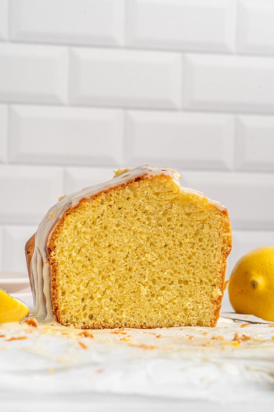 A sliced loaf of lemon drizzle cake with a light glaze on top is displayed against a white tiled background. Lemons are placed nearby, and crumbs are scattered around the cake.