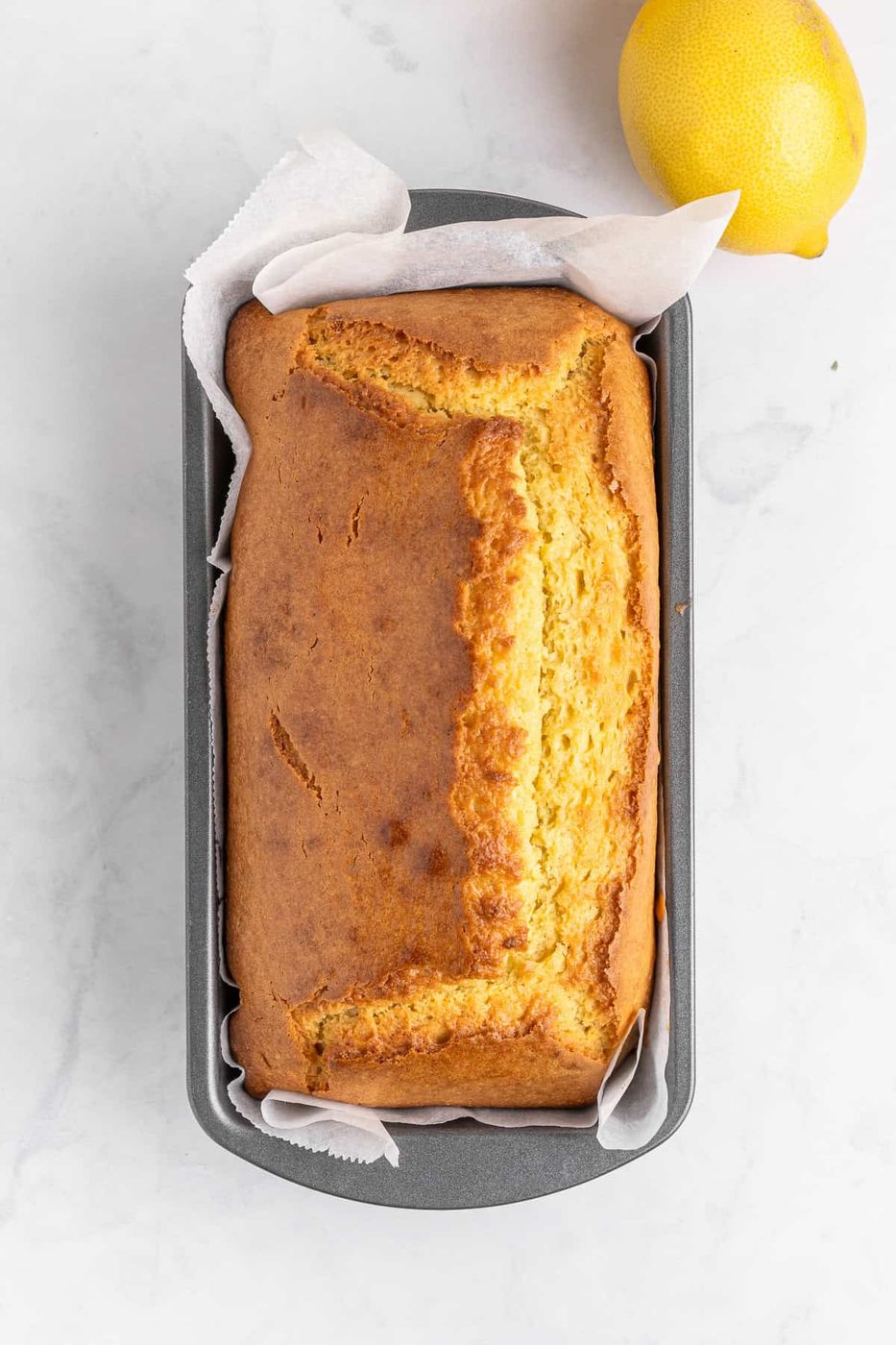 A loaf of golden-brown lemon bread in a parchment-lined loaf pan on a marble surface. A whole lemon lies beside the pan.