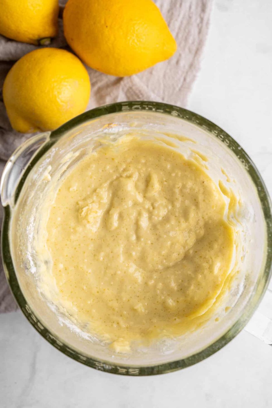 A glass mixing bowl filled with creamy lemon batter on a white countertop. Three whole lemons lie on a beige cloth beside the bowl.