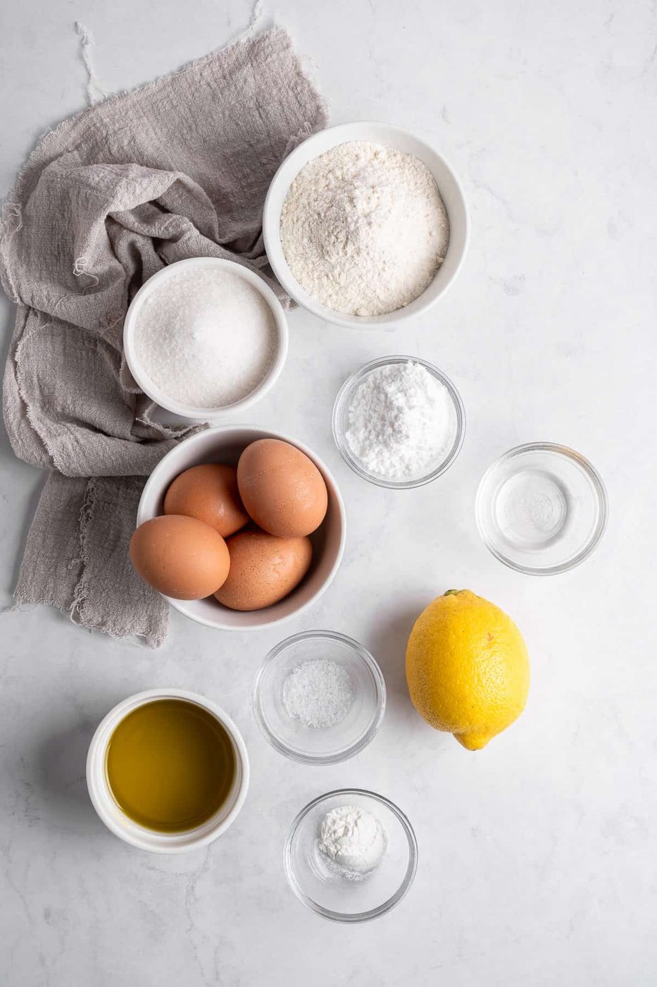 A flat lay of baking ingredients on a marble surface, including flour, sugar, eggs, a lemon, olive oil, baking powder, salt, and water. A gray cloth napkin is placed alongside the ingredients.