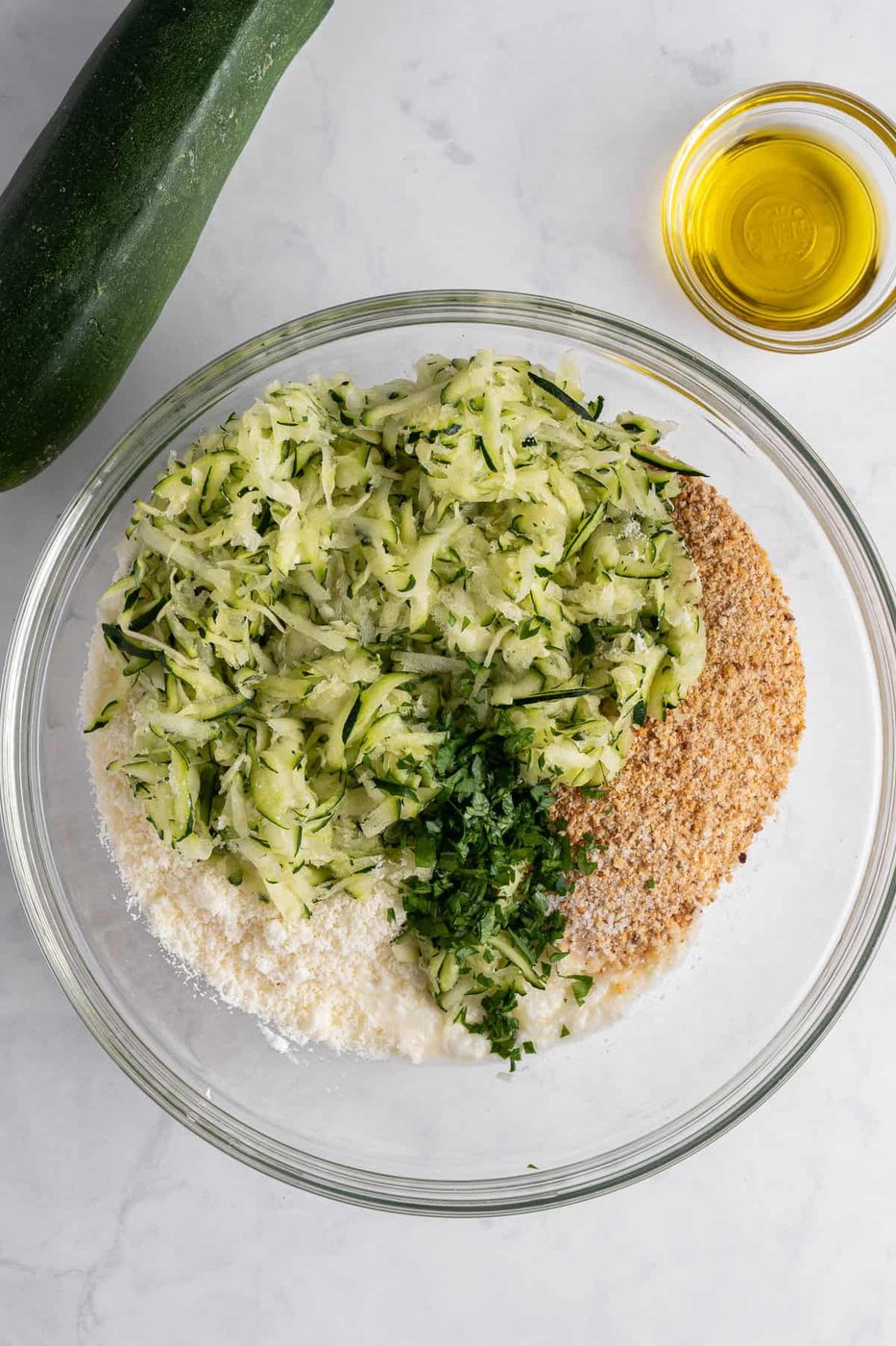 A bowl with grated zucchini, breadcrumbs, parmesan cheese, and chopped herbs on a marble surface. A whole zucchini and a small bowl of olive oil are next to it.