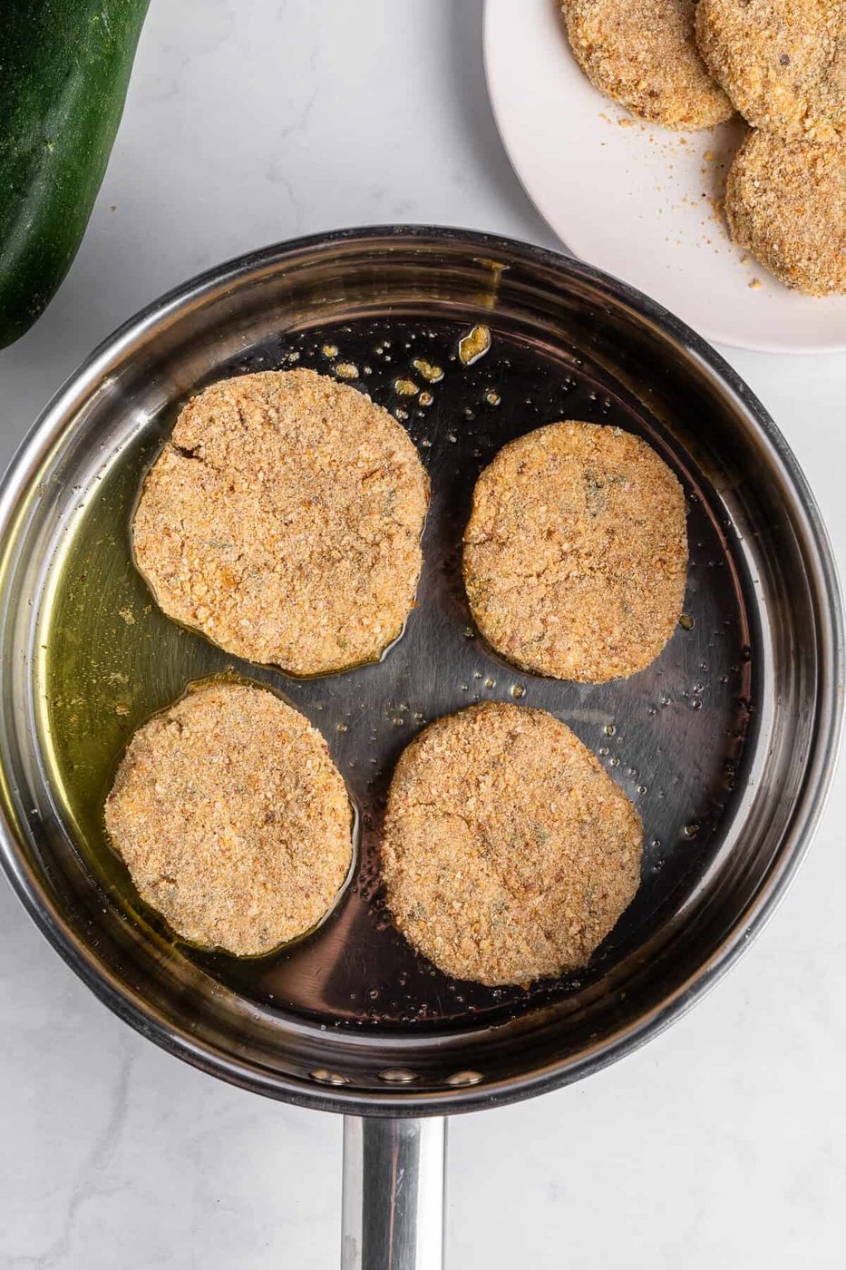 Four breaded zucchini slices are frying in a stainless steel pan with oil. A plate with more breaded slices is visible in the background.