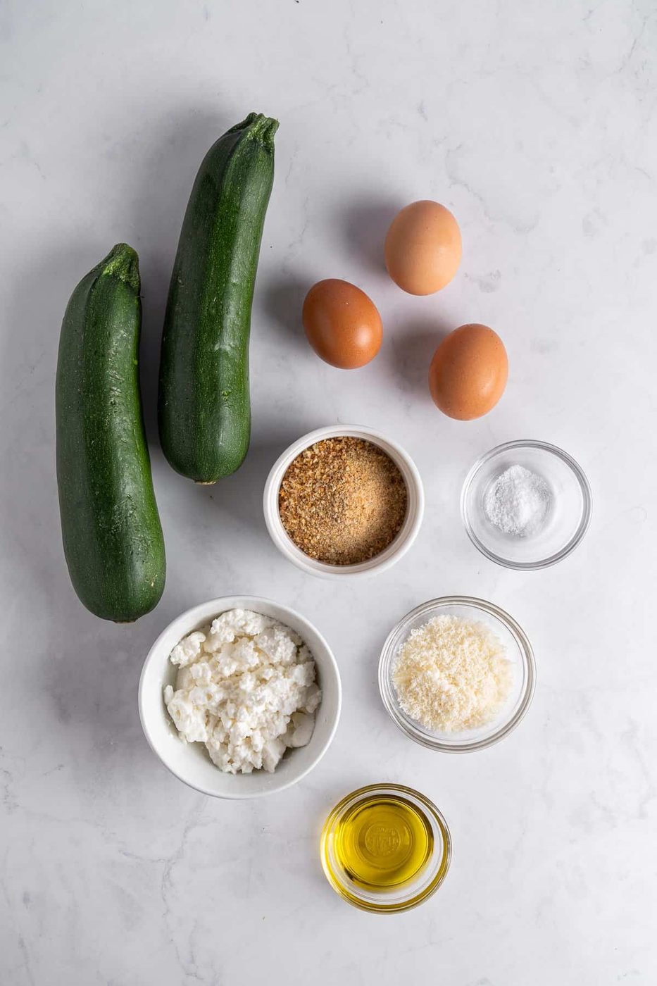 Ingredients arranged on a marble surface: two zucchinis, three eggs, breadcrumbs in a bowl, salt in a small bowl, ricotta cheese in a bowl, grated Parmesan in a bowl, and a bowl of olive oil.