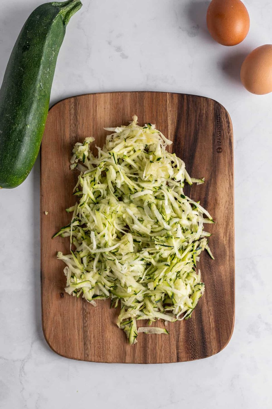 A wooden cutting board holds a pile of shredded zucchini. A whole zucchini is placed beside the board, along with two brown eggs on a white marble surface.