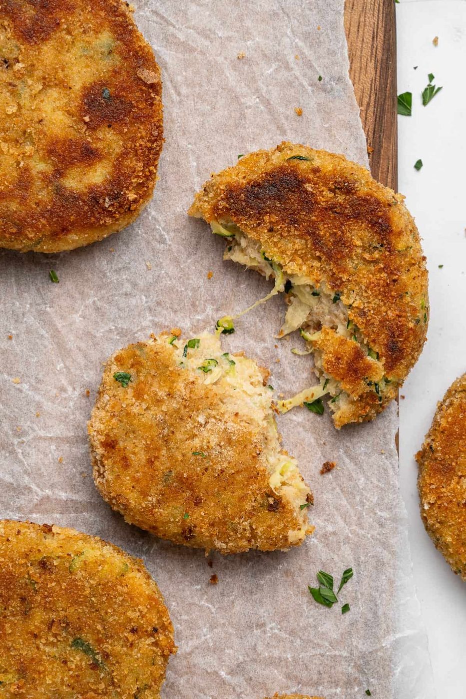 Top view of breaded and fried patties on parchment paper. One patty is broken in half, revealing a creamy filling with herbs. Other patties are whole and garnished with chopped parsley, adding a fresh green touch.