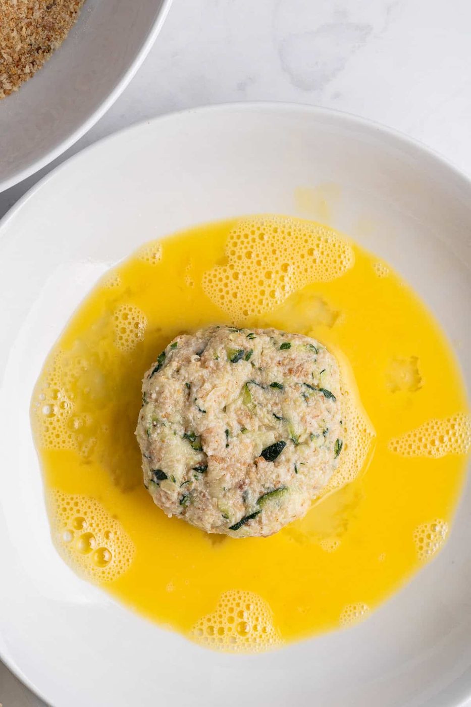 A plate holds a mixture of minced meat and herbs in a pool of beaten egg. The egg yolk and whites are mixed, creating a yellow liquid around the meat, which appears partially formed into a patty.