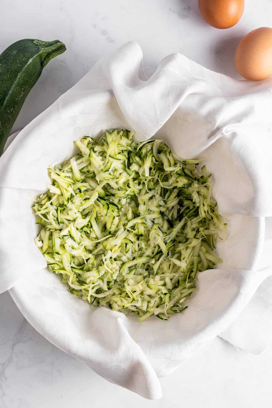 A bowl lined with a white cloth holds grated zucchini on a marble countertop. A whole zucchini and two eggs are positioned nearby.