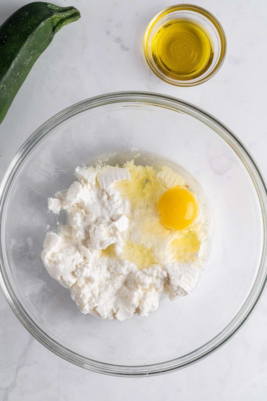A glass bowl with ricotta cheese and a cracked egg on a marble countertop. Nearby, a small glass bowl contains olive oil, and a zucchini is partly visible.