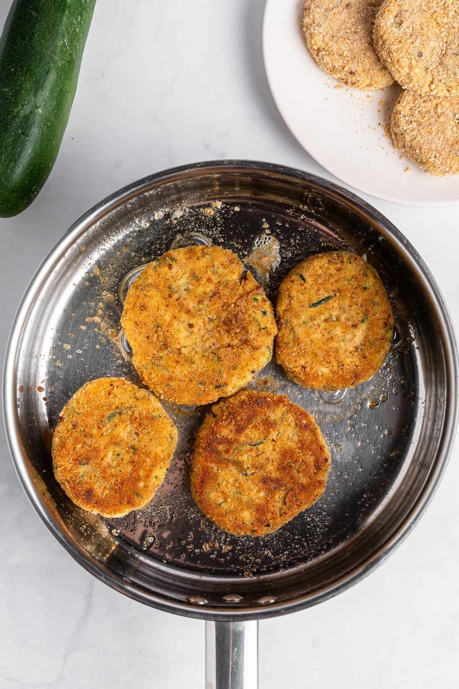 Four golden-brown zucchini fritters are frying in a stainless steel pan. A zucchini is nearby, and a plate with breadcrumbs and uncooked fritters is in the background on a white marble surface.