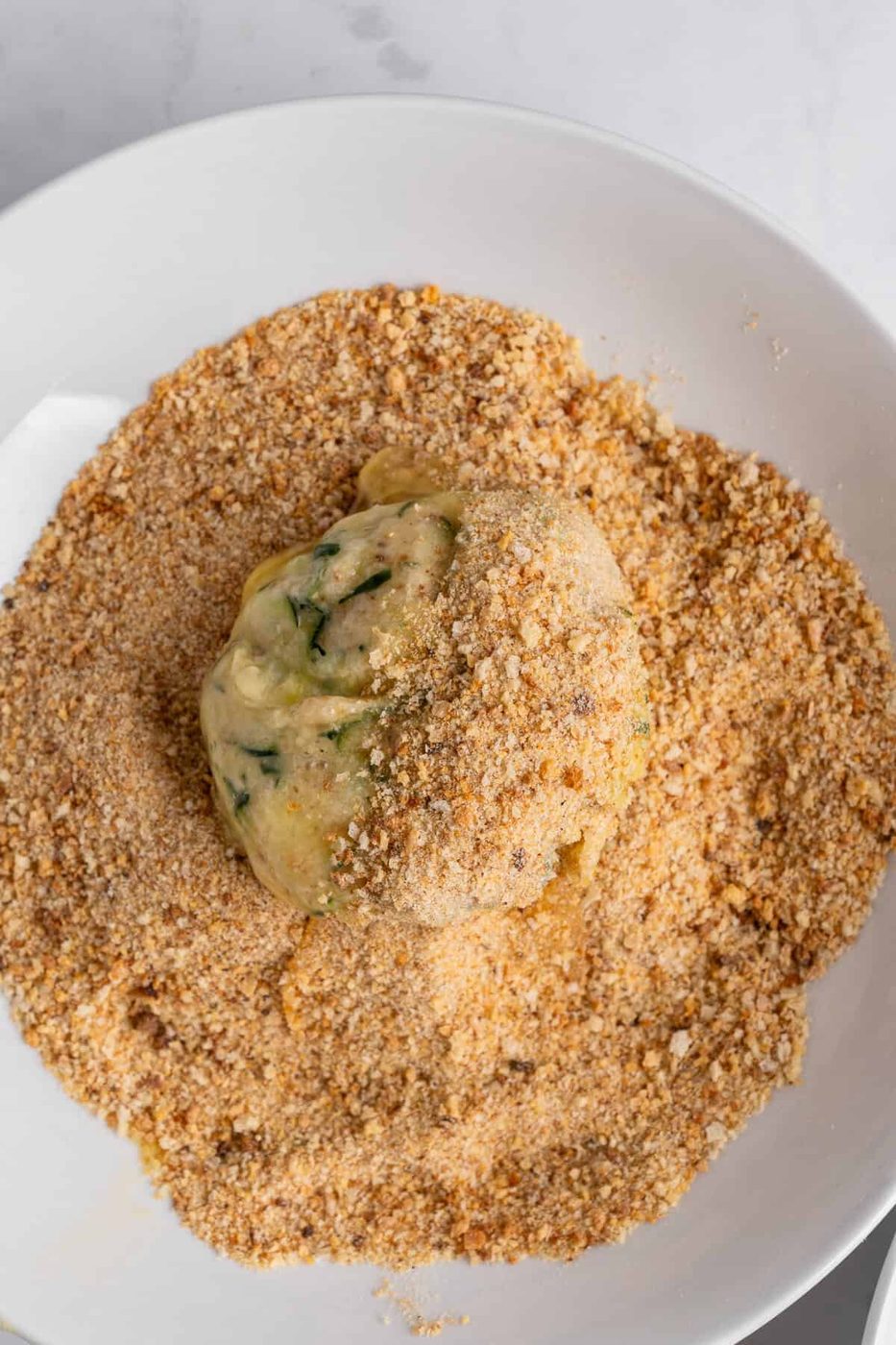A ball of uncooked dough, possibly containing herbs, is partially coated in a layer of breadcrumbs on a white plate atop a marble surface.
