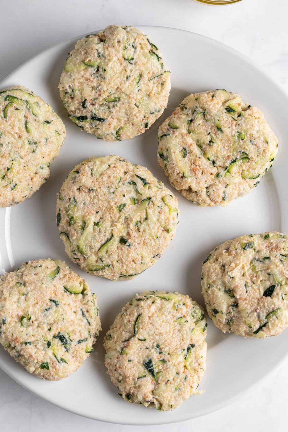 A white plate with six uncooked, round patties made of grated zucchini and breadcrumbs. The patties have a speckled texture with visible green bits of zucchini throughout.