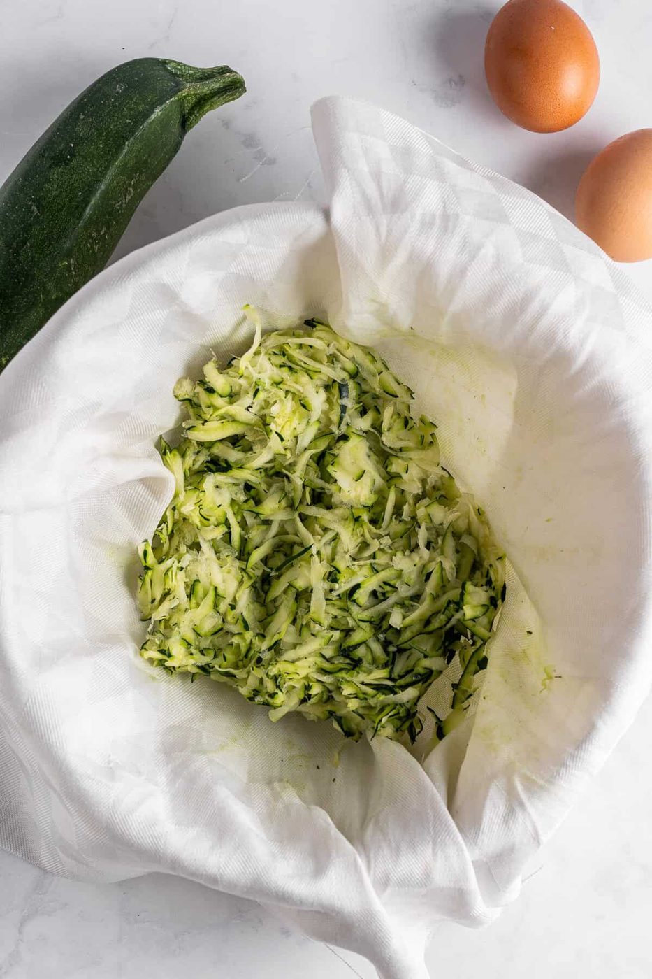 A pile of shredded zucchini rests on a white cloth, surrounded by a whole zucchini and two brown eggs on a marble countertop.