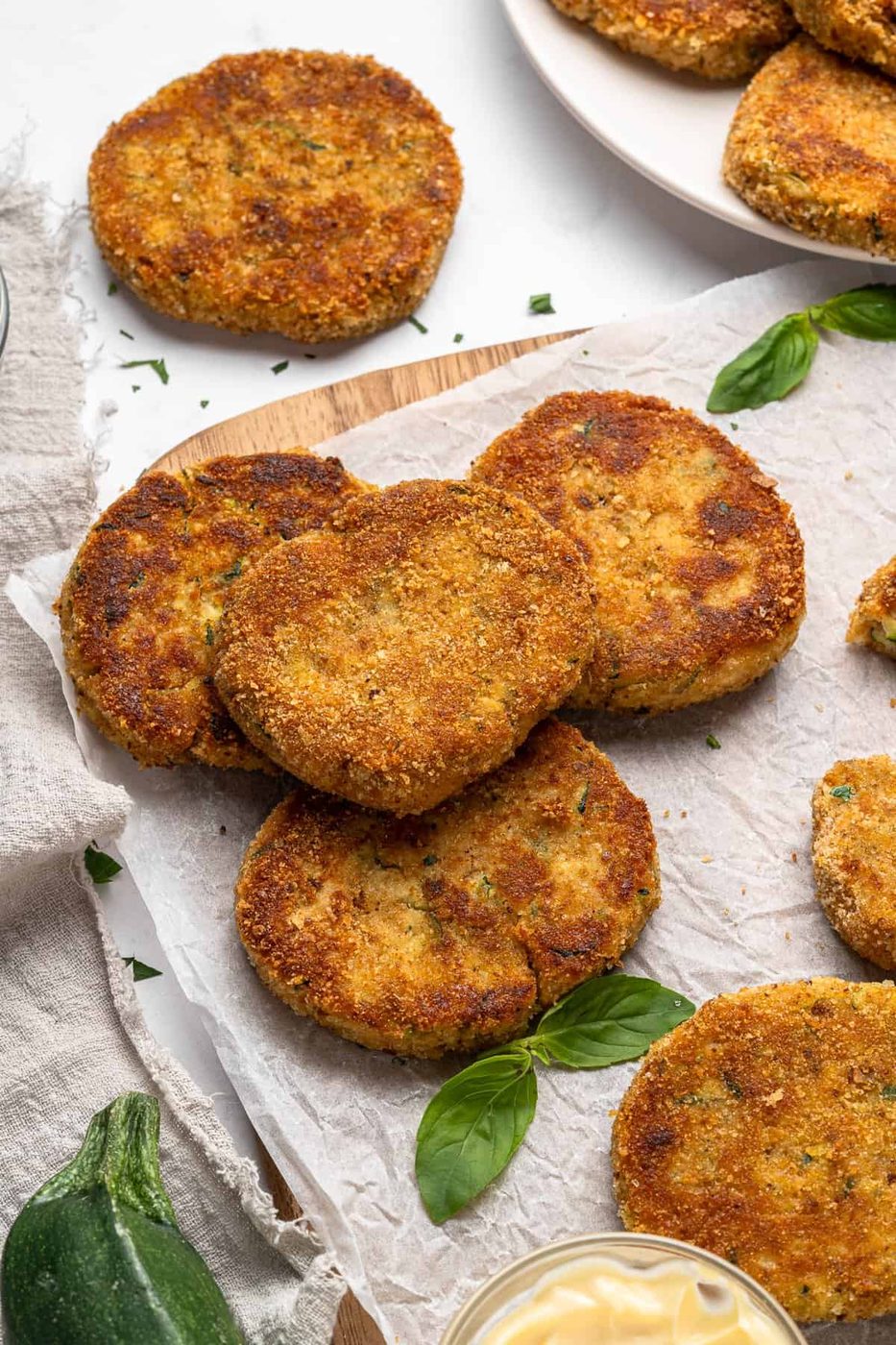 Golden-brown, zucchini cakes are stacked on parchment paper with fresh basil leaves. A small bowl of dipping sauce is nearby, and more patties are displayed on a plate in the background. A zucchini is partially visible.