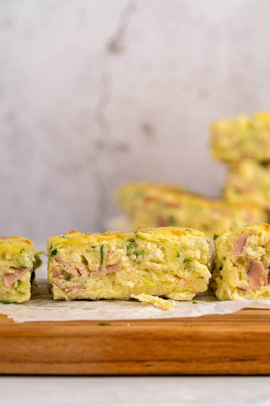 Close-up of a slice of savory egg bake with visible pieces of ham and green herbs, resting on parchment paper atop a wooden board. More pieces are blurred in the background.