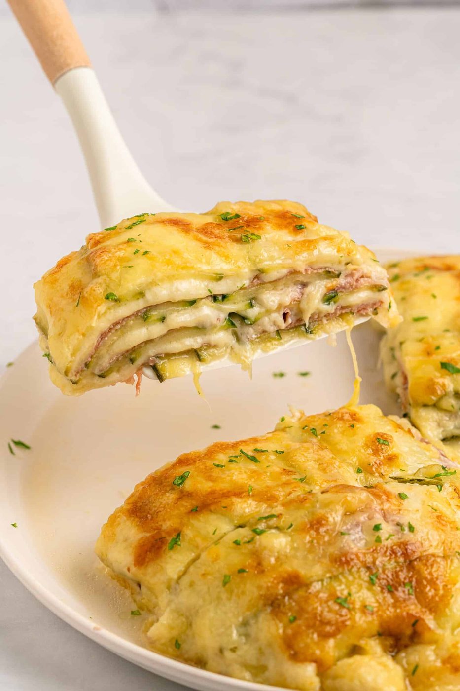 A cheesy slice of baked lasagna with herbs is lifted from a white plate, showing layers of pasta, melted cheese, and filling. Another portion remains on the plate in the background.