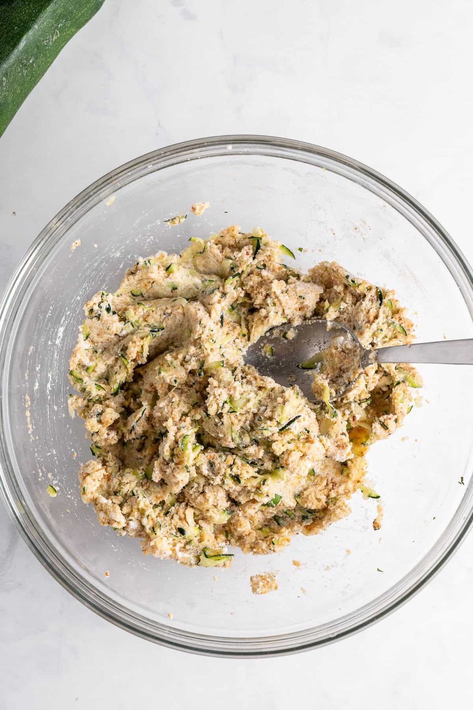A glass bowl filled with a mixture of grated zucchini and dough, with a metal spoon resting inside. The bowl is placed on a white marble surface, and part of a zucchini is visible in the top left corner.