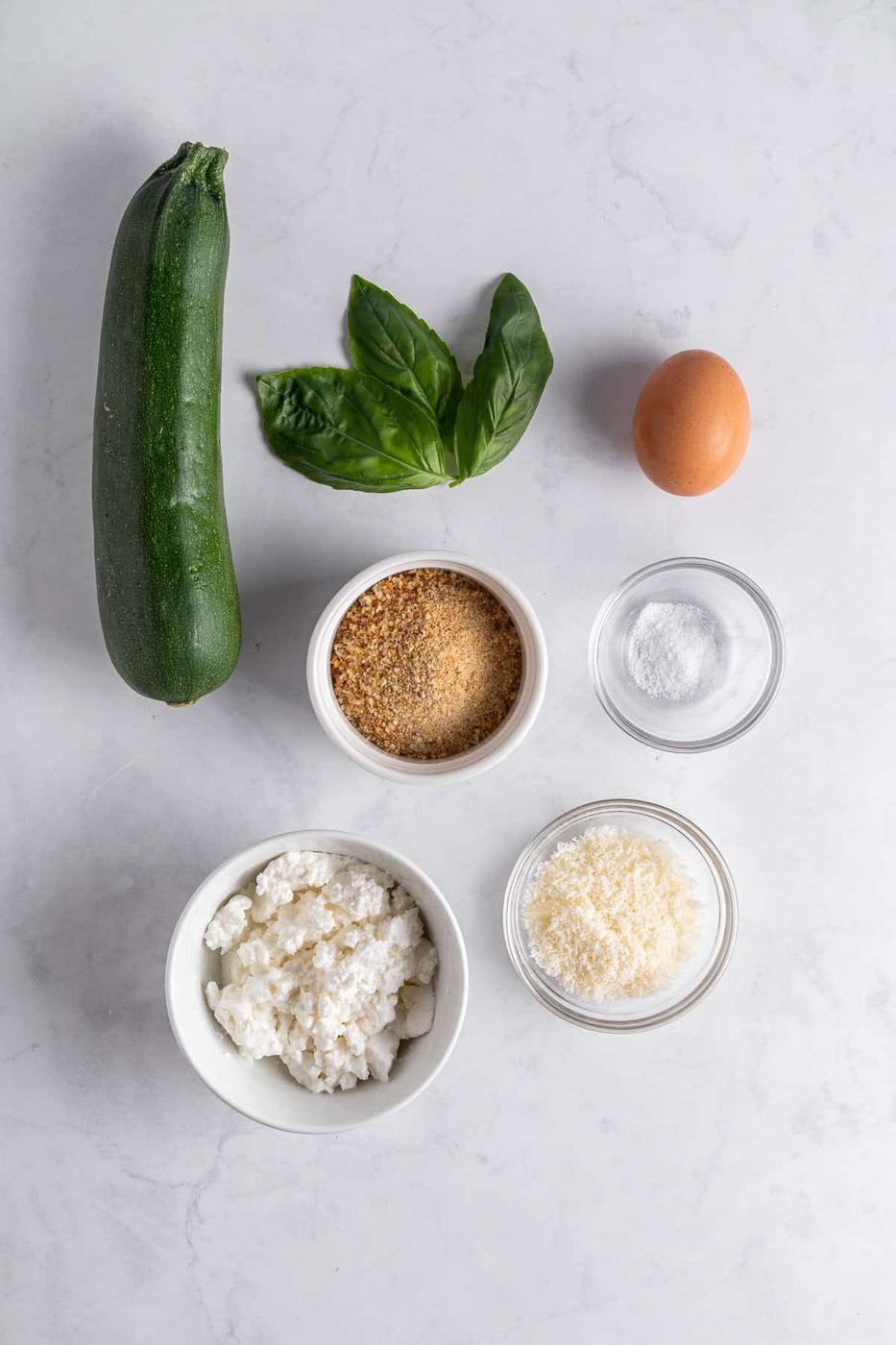 A zucchini, fresh basil leaves, an egg, and small bowls of breadcrumbs, salt, ricotta cheese, and grated Parmesan are arranged on a light marble surface.