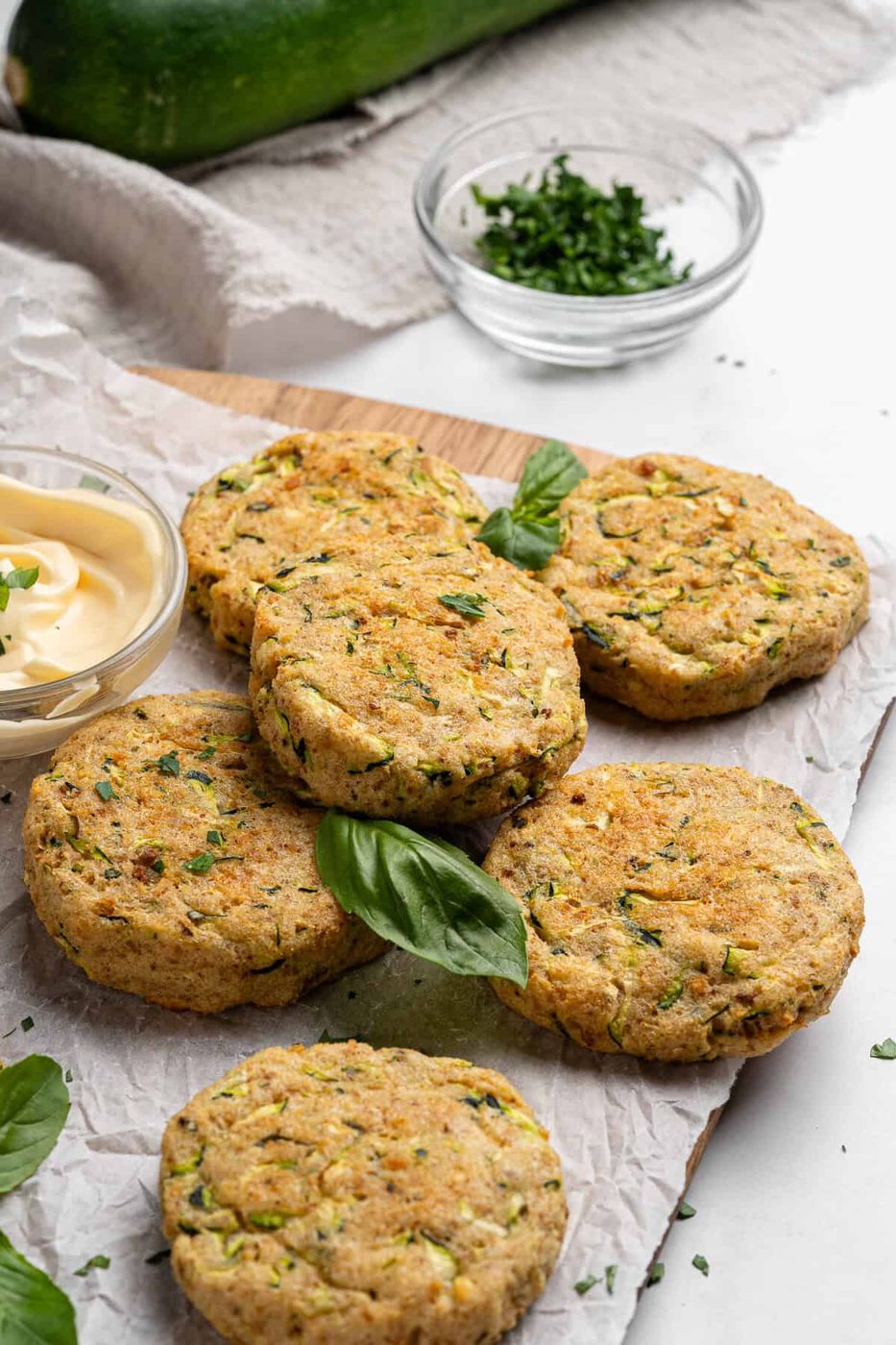Five zucchini fritters are arranged on a piece of parchment paper, garnished with fresh basil leaves. A small glass bowl of chopped herbs and a bowl of mayonnaise are nearby. A whole zucchini and a linen cloth are in the background.