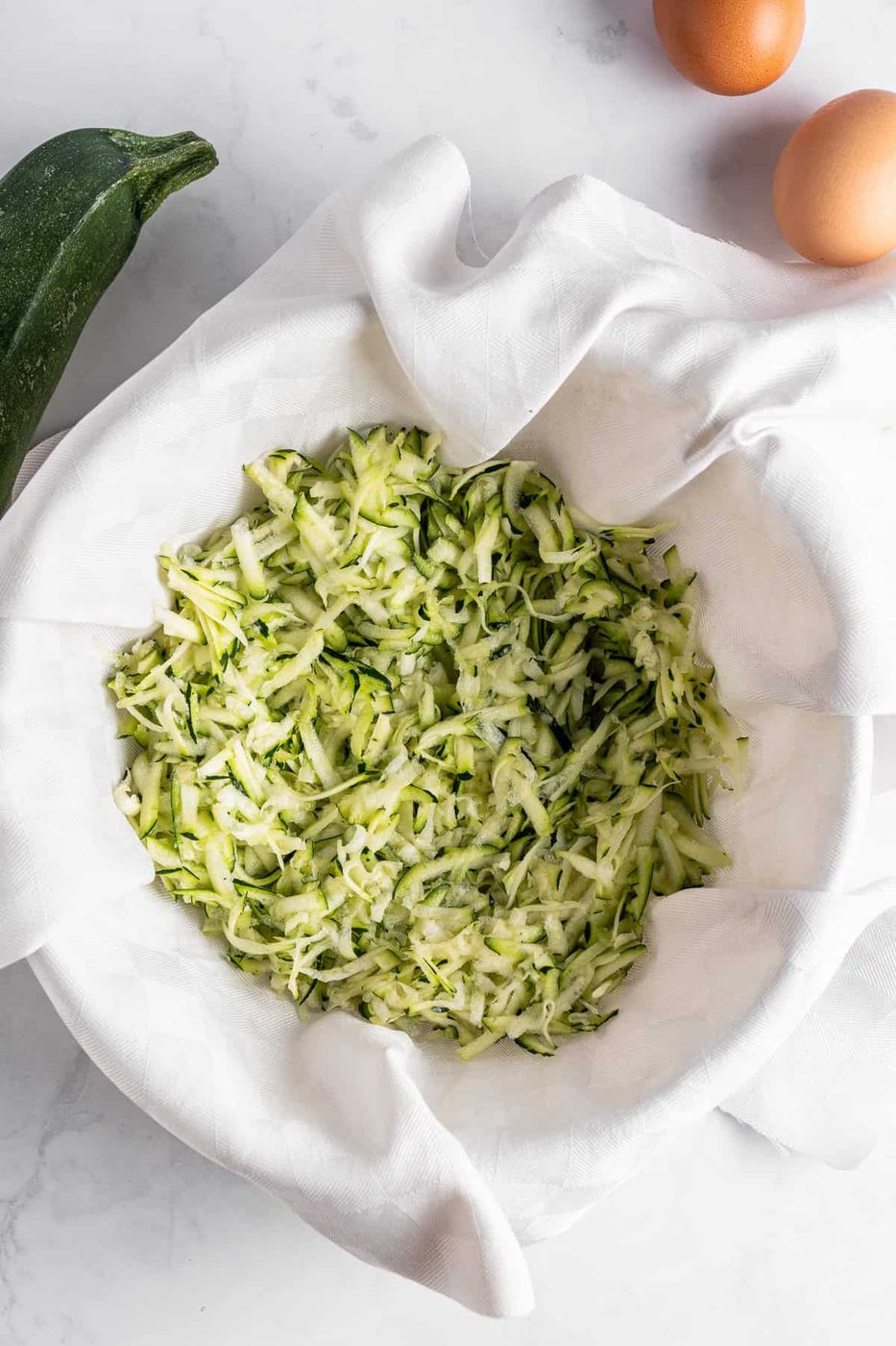 Shredded zucchini in a white cloth-lined bowl on a marble counter, with a whole zucchini and two eggs nearby.