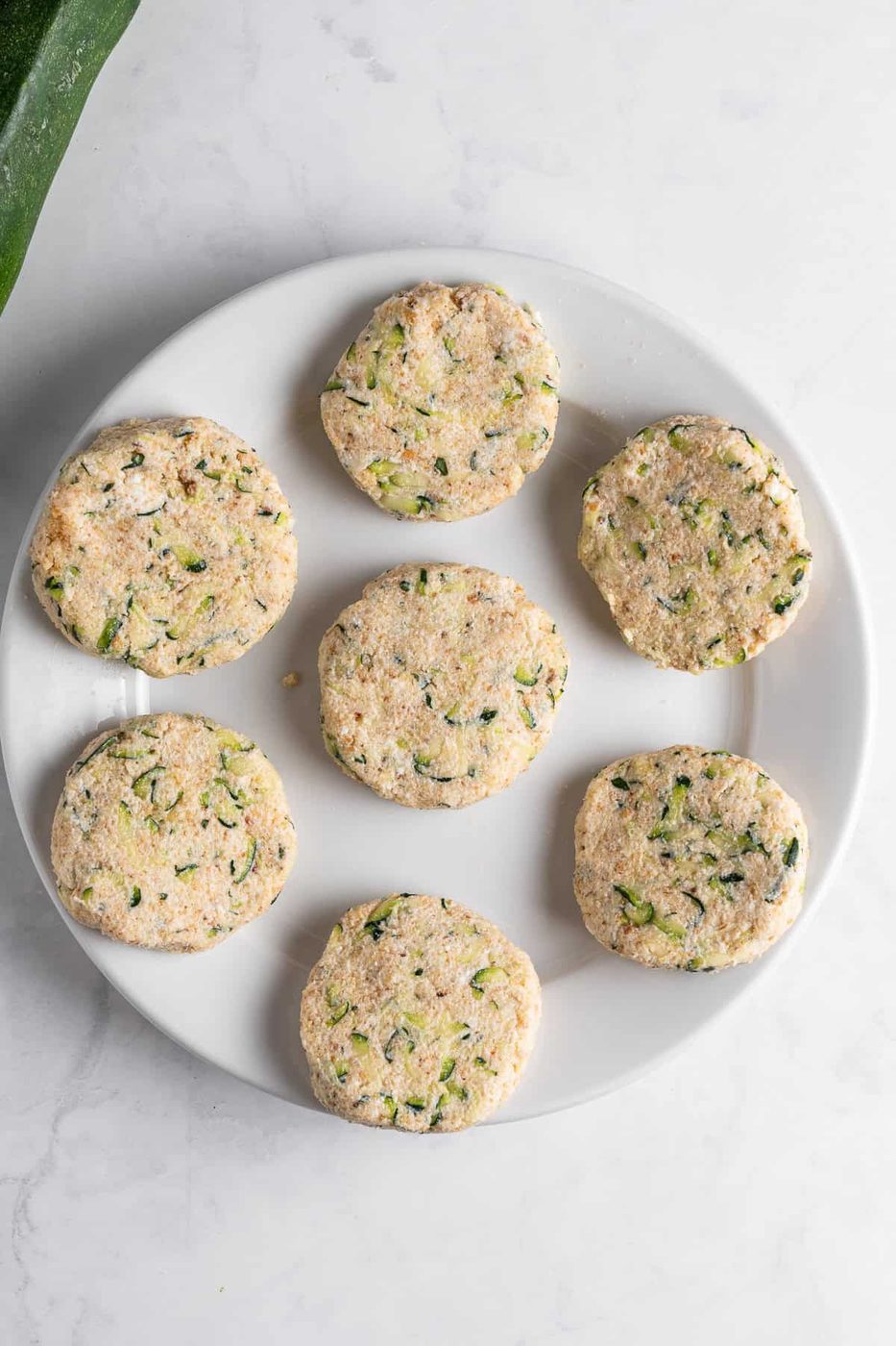 A white plate with seven uncooked, round zucchini fritters, featuring visible zucchini and herbs, placed on a marble countertop.