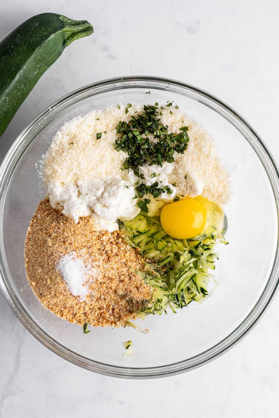 A glass bowl containing grated zucchini, an egg, breadcrumbs, grated cheese, chopped herbs, and a bit of white ricotta cheese on a marble surface. A whole zucchini lies beside the bowl.