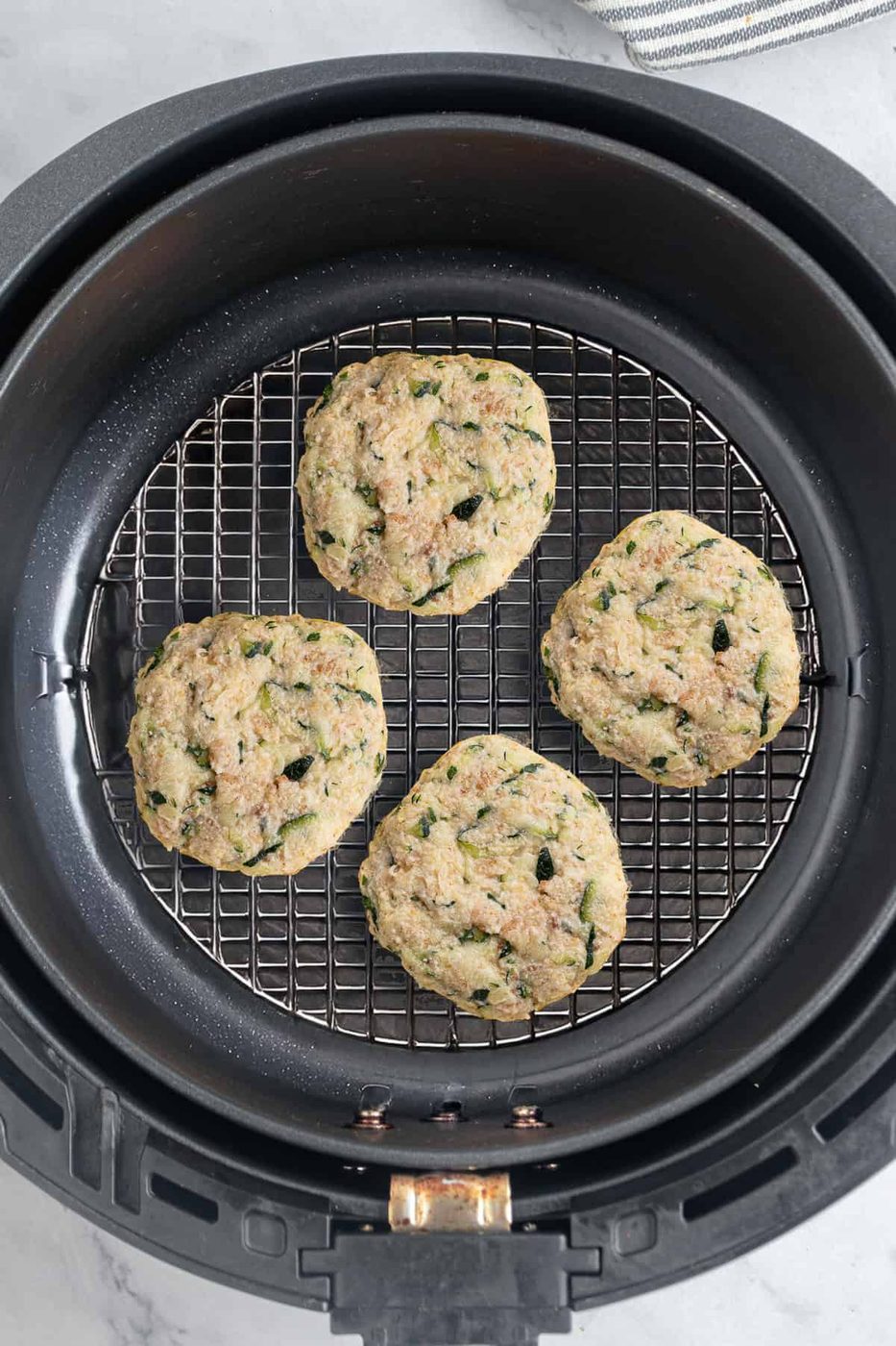 Four zucchini fritters are arranged on a wire rack inside an air fryer basket. They have a golden-brown appearance and are speckled with green zucchini pieces. The background shows part of a gray and white striped cloth.