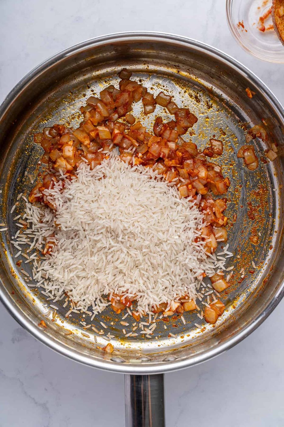 A stainless steel pan on a marble surface contains sautéed onions and a heap of uncooked white rice. The onions appear to be seasoned with spices, giving them a reddish-brown color.