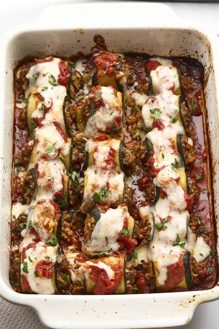 A baked dish featuring zucchini roll-ups filled with a savory mixture, topped with melted cheese and tomato sauce. The roll-ups are garnished with chopped herbs, all within a rectangular baking dish.