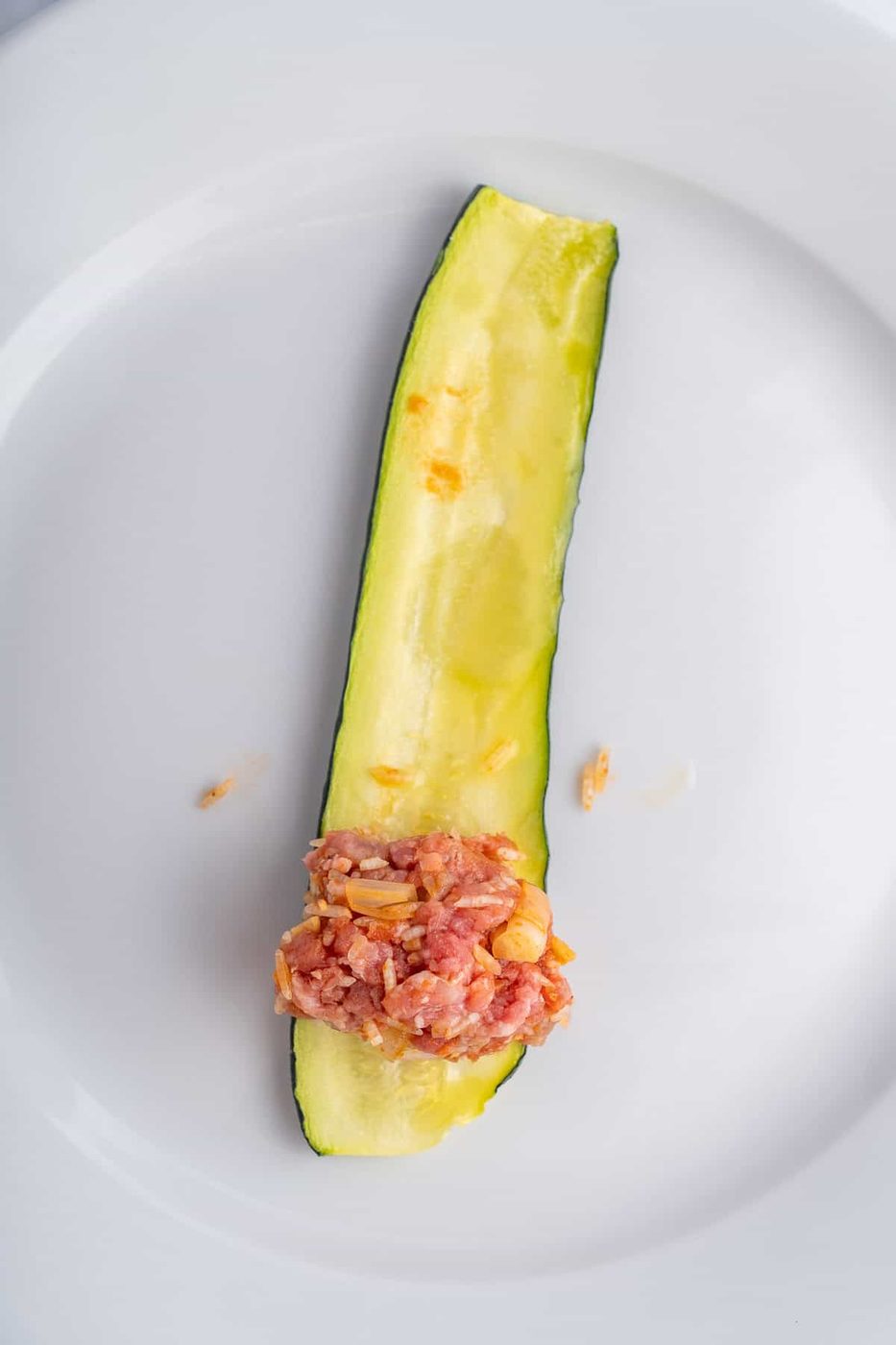 A single slice of zucchini on a white plate, hollowed out and topped with a small portion of minced meat mixed with onions.