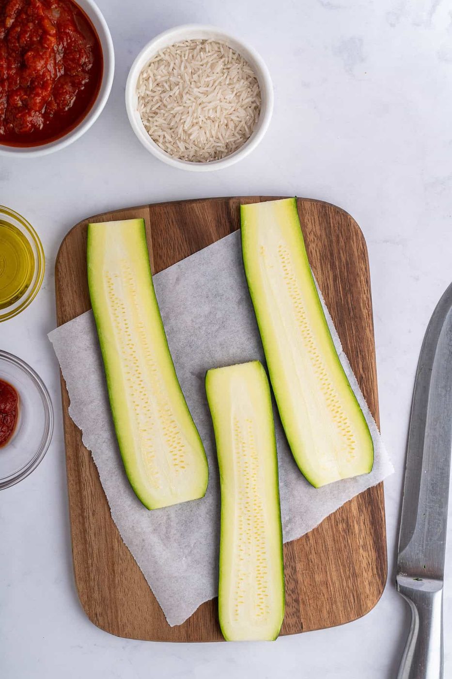 Sliced zucchini on a wooden board with parchment paper, surrounded by bowls of rice, tomato sauce, and olive oil. A knife is visible on the right side.