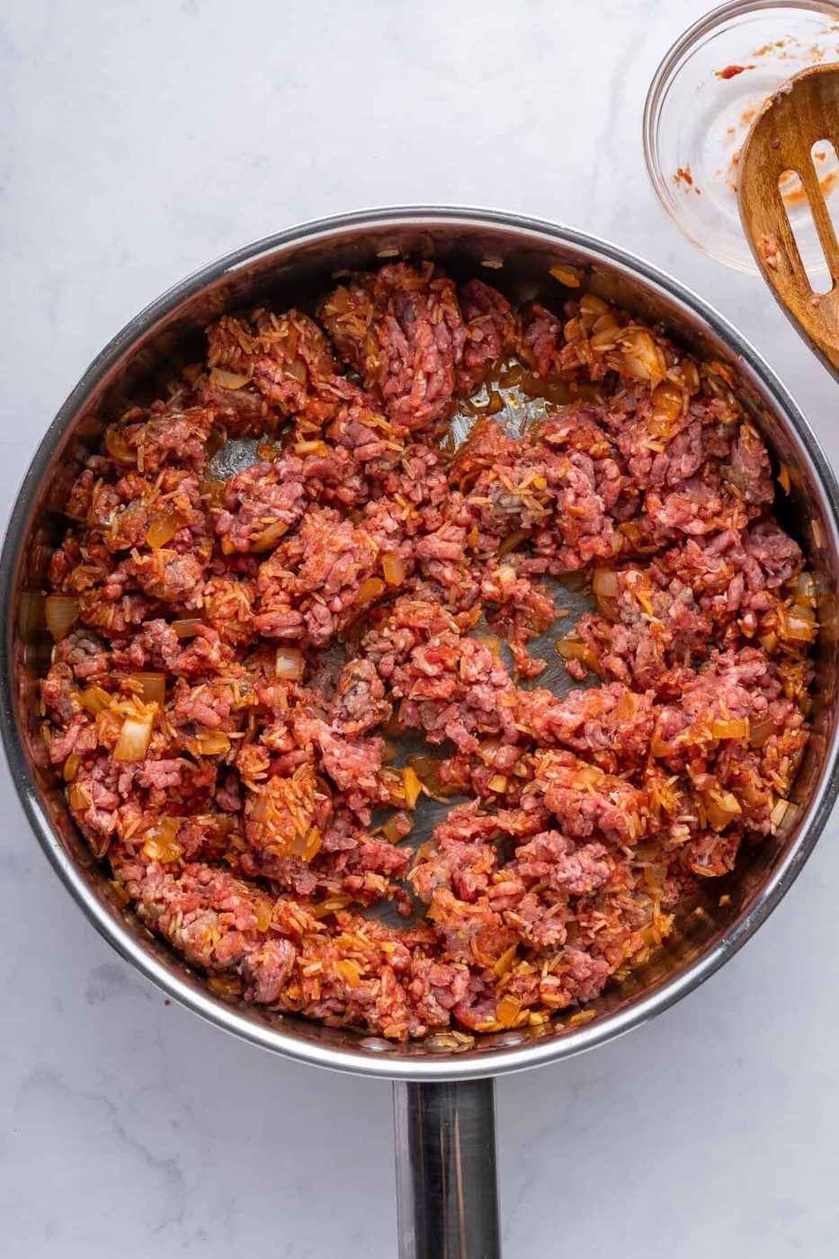 A frying pan filled with minced meat cooking on a stovetop. The meat is partially cooked, mixed with chopped onions and other ingredients, and has a reddish hue. The background is a light, textured surface. A wooden spoon rests nearby.