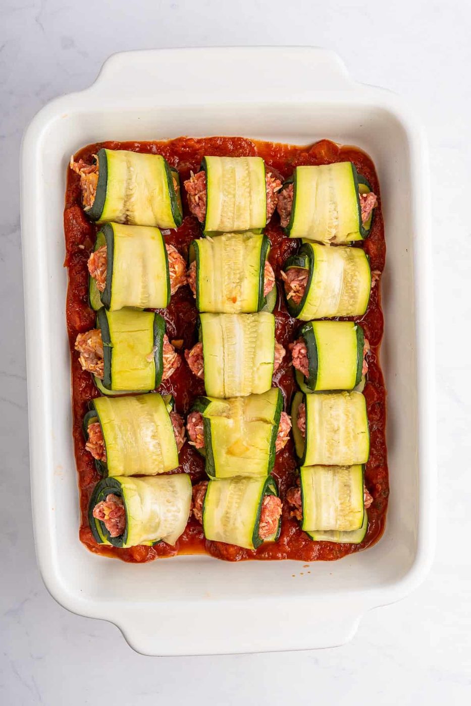 A white baking dish filled with zucchini roll-ups on a bed of tomato sauce. The roll-ups are made with thin zucchini slices wrapped around a filling, possibly meat or a plant-based alternative. The dish sits on a white marble surface.