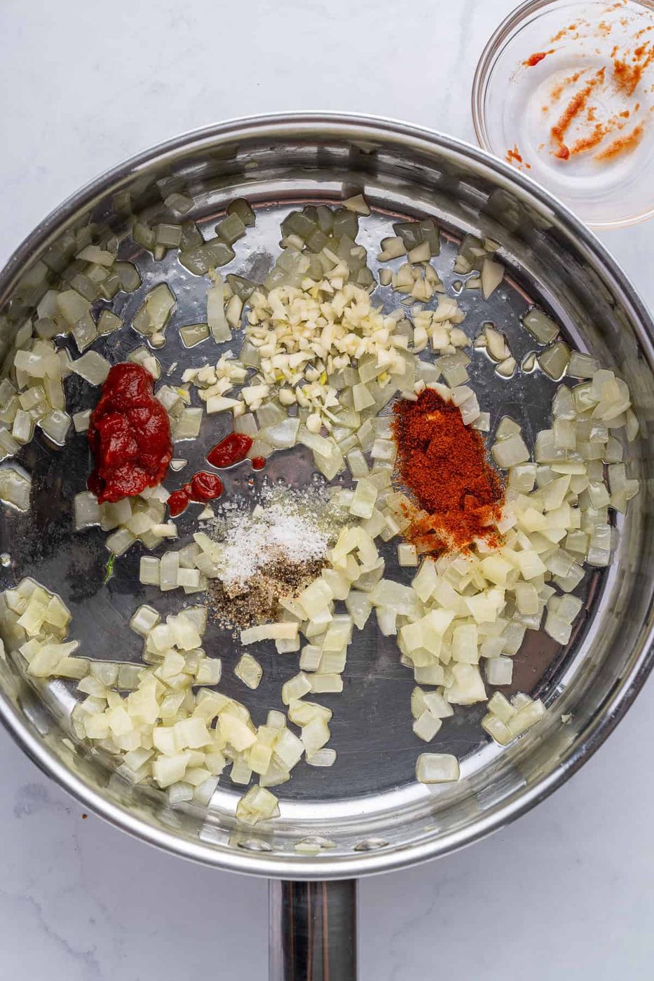 A stainless steel pan with sautéed onions, minced garlic, tomato paste, paprika, and seasoning. A small glass bowl with spices is nearby on a white marble surface.