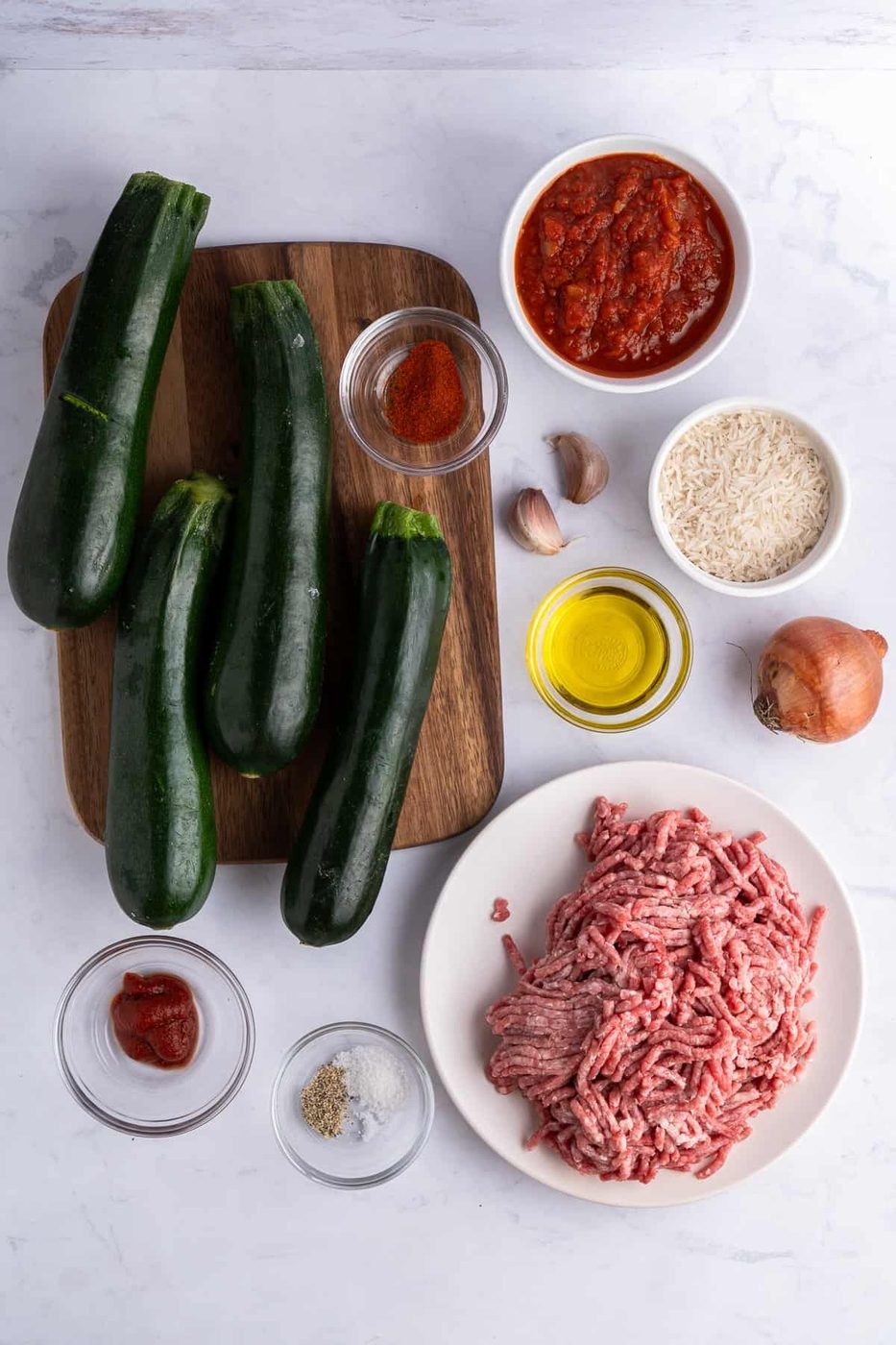Ingredients for a dish arranged on a marble surface: several zucchinis on a wooden board, bowls of tomato sauce, rice, ground meat, spices, olive oil, and small bowls containing tomato paste, salt, pepper, garlic, and a whole onion.