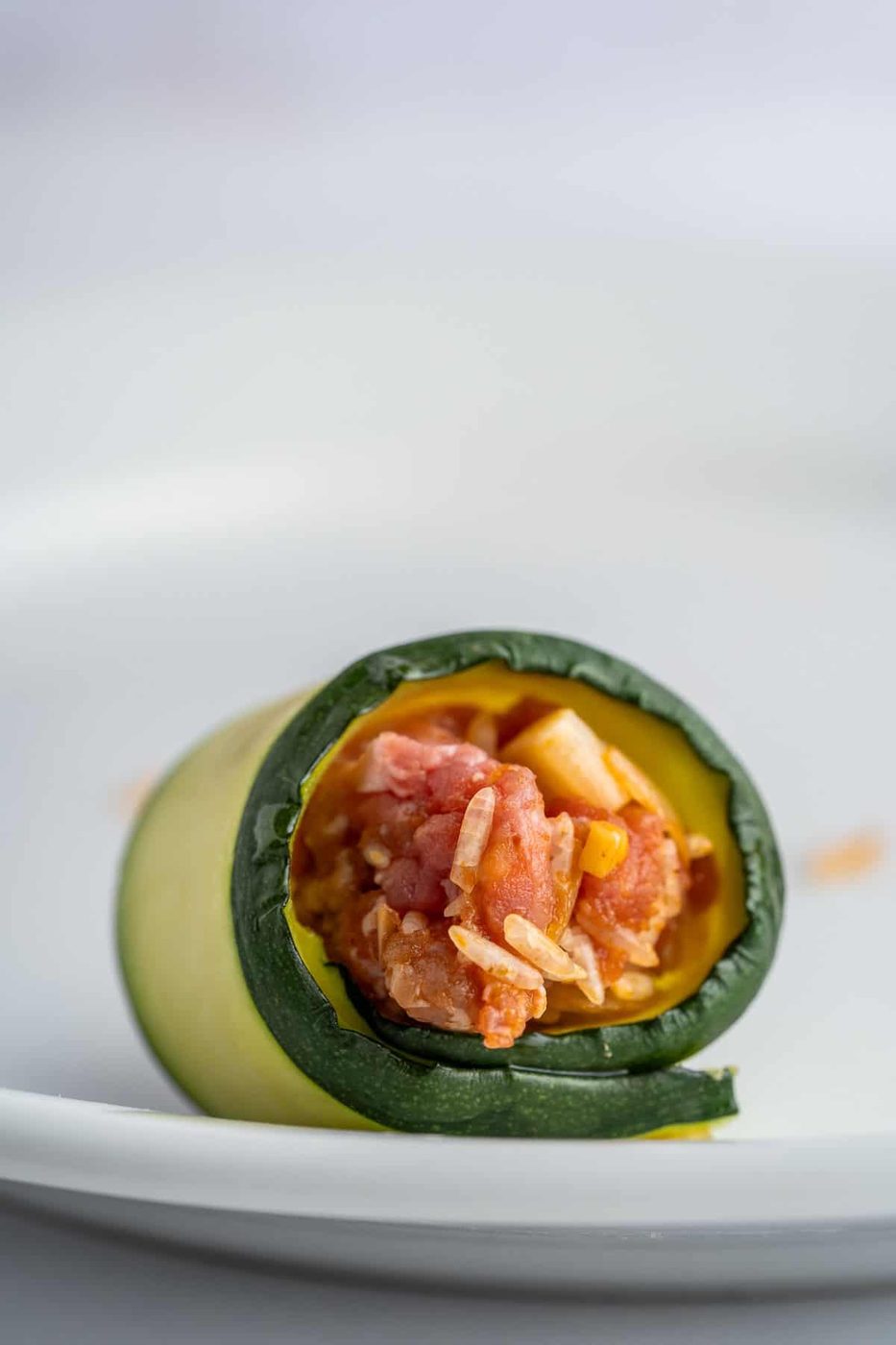 Close-up of a zucchini roll filled with a mixture of diced tomatoes, corn, and shredded cheese, placed on a white plate.