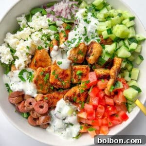 An Easy Greek Bowl filled with grilled chicken, diced cucumber, tomato, red onion, olives, feta cheese, and a drizzle of creamy white dressing, all garnished with chopped parsley.
