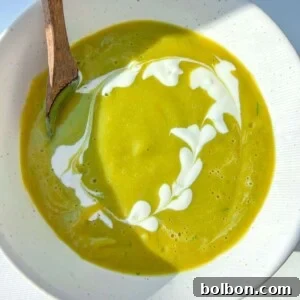 A bowl of Healthy Cream of Broccoli Soup with a swirl of white cream forming a heart-shaped pattern, served with a wooden spoon on the side. Bright natural light casts soft shadows across the bowl.