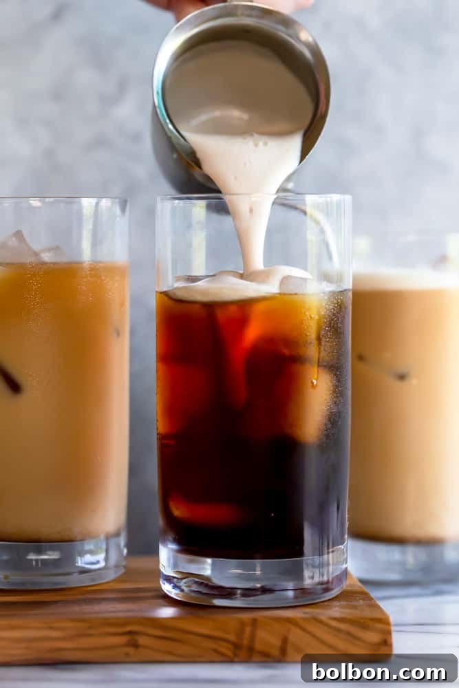 An artfully arranged scene featuring a glass of homemade Pumpkin Cream Cold Brew next to an espresso machine, surrounded by fall leaves, small squash, and a coffee mug, suggesting a home barista setup for autumn beverages.