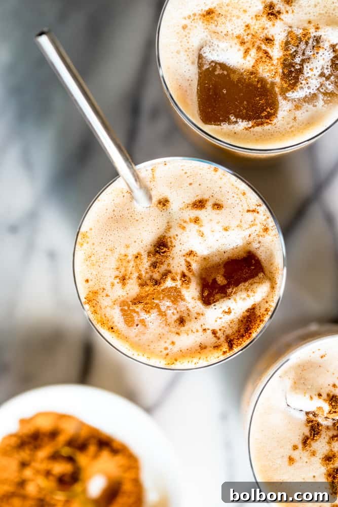 A hand gently pouring a cascade of light, frothy pumpkin cream into a clear glass filled with ice and dark cold brew coffee, demonstrating the final assembly of the homemade beverage.