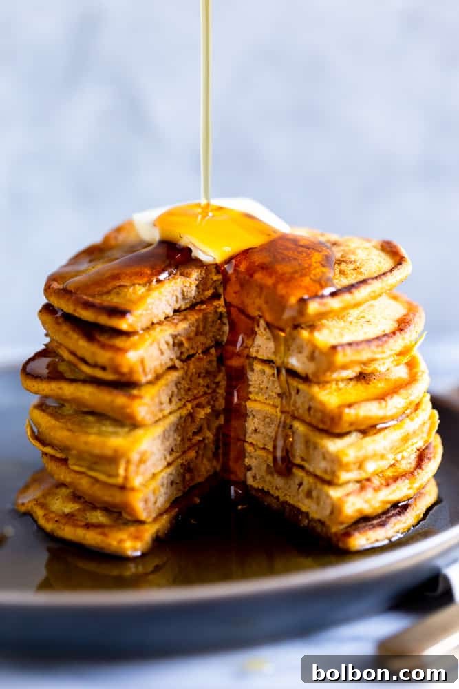 Syrup pouring generously onto a tall stack of golden brown, fluffy pumpkin pancakes, with a slice already cut out, ready to be enjoyed.