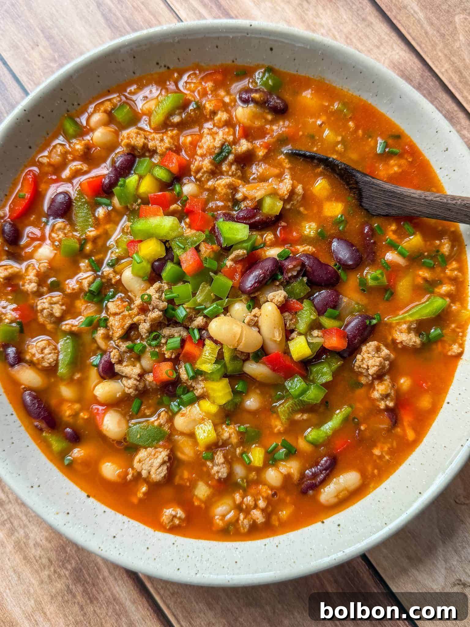 Guilt-Free Turkey Chili 3 A close-up of healthy turkey chili in a rustic bowl, showcasing kidney beans, cannellini beans, ground turkey, and colorful bell peppers, topped with fresh chives, with a wooden spoon for serving.