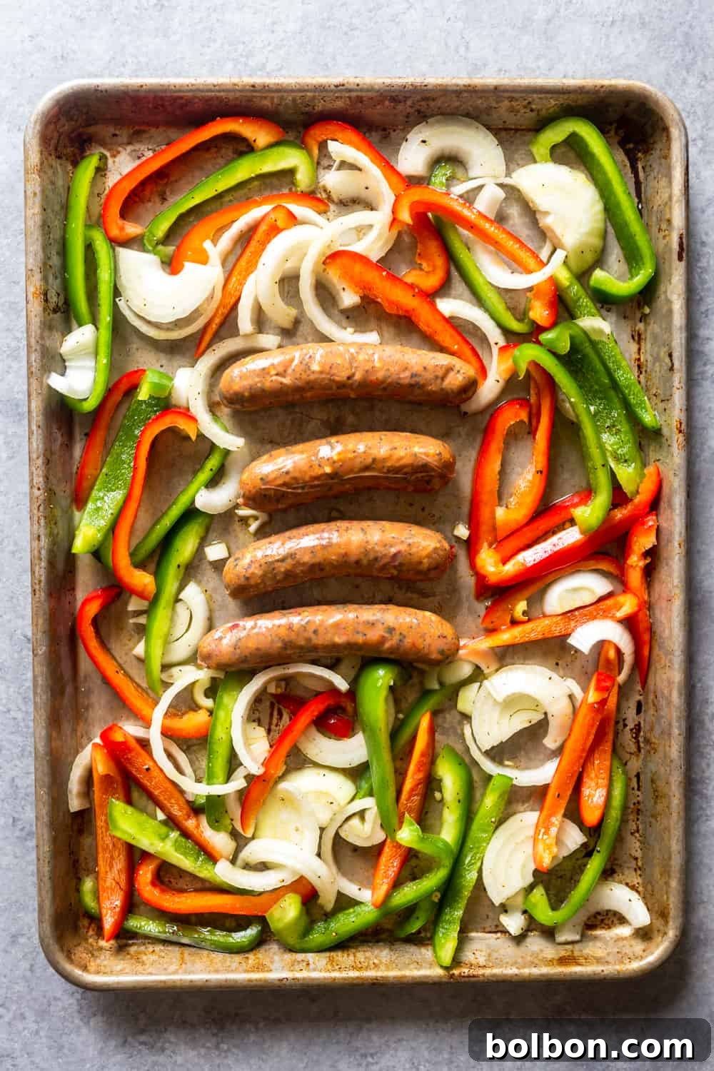 Preparing Sheet Pan Sausage and Peppers Raw bell peppers, sliced onions, and sausages arranged on a baking sheet, pre-oven.