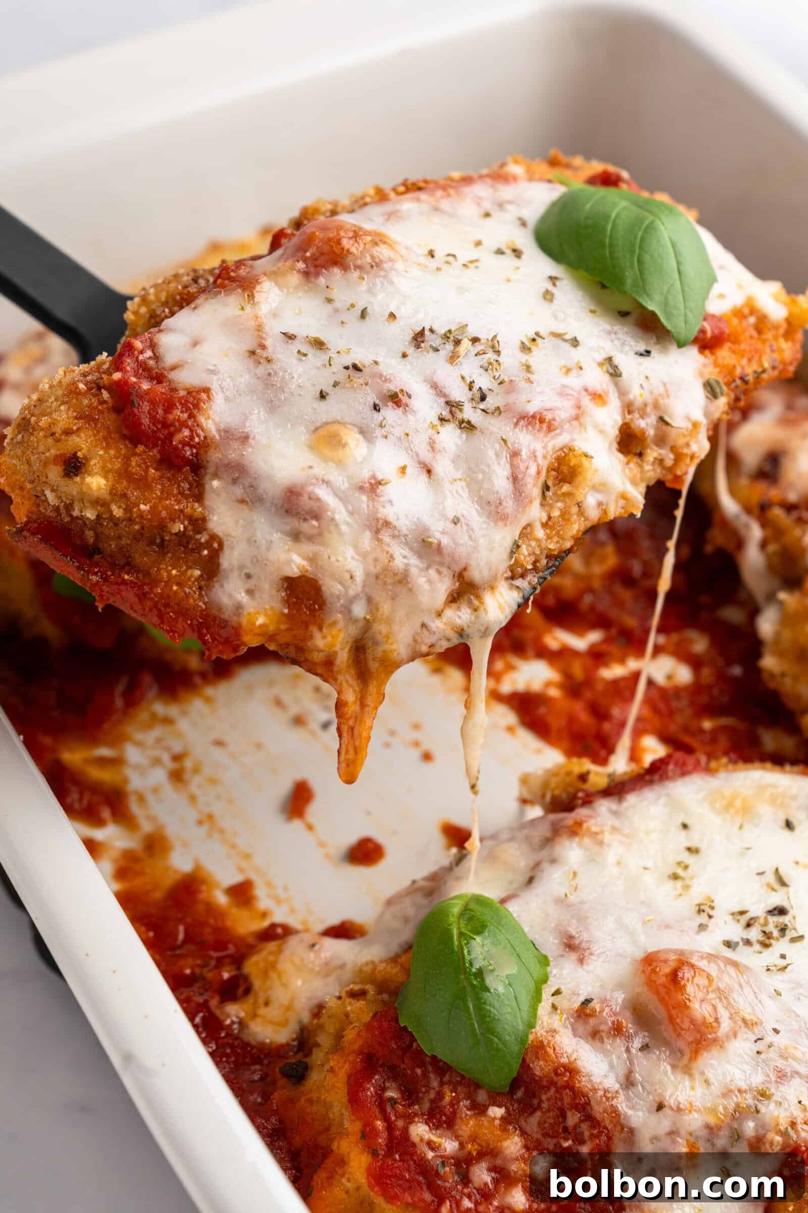 Homestyle Baked Chicken Parmigiana 2 A close-up of a dish of chicken parmigiana being served. The piece of chicken is topped with melted mozzarella cheese, tomato sauce, and garnished with fresh basil. It's being lifted from a white baking dish with more chicken pieces inside.