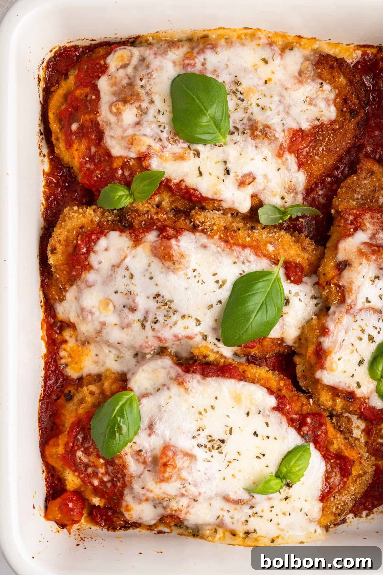 Homestyle Baked Chicken Parmigiana 11 A baking dish with four pieces of chicken parmigiana topped with melted cheese and tomato sauce, garnished with fresh basil leaves. The dish is golden and crispy around the edges.
