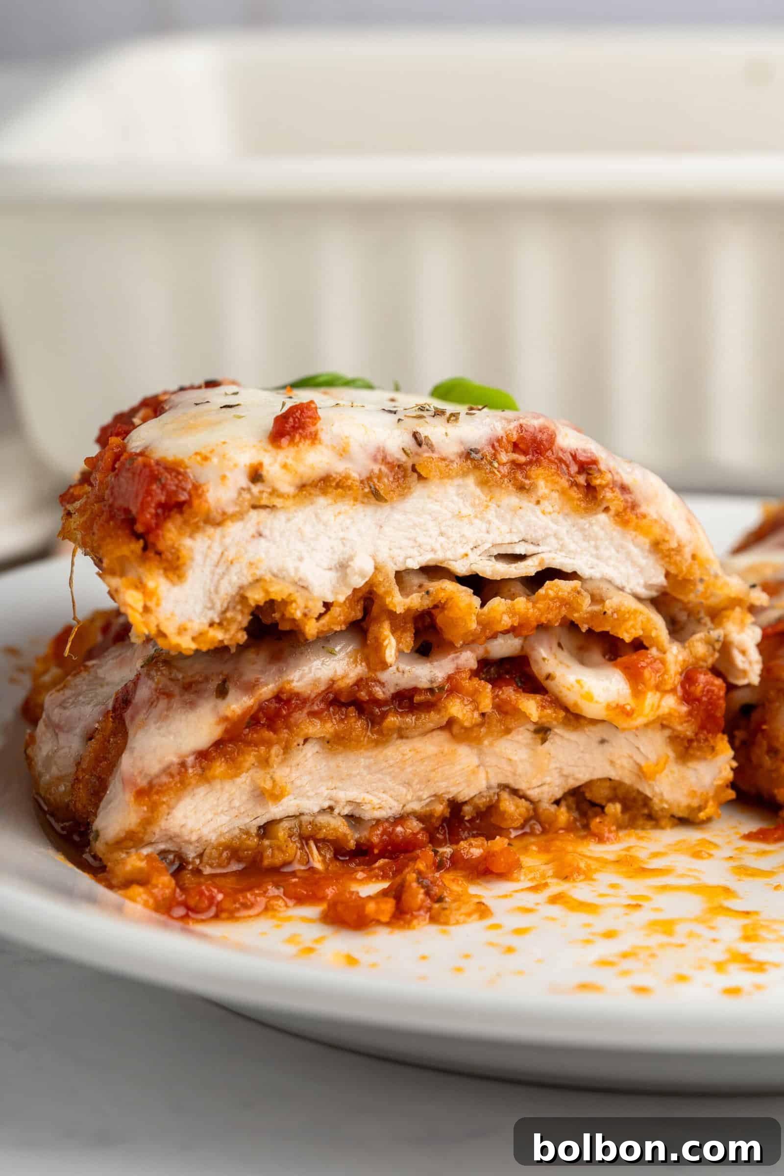 Homestyle Baked Chicken Parmigiana 12 Close-up of a sliced chicken Parmesan dish on a white plate. Layers of crispy chicken, melted cheese, and tomato sauce are visible, with some fresh basil on top. A baking dish is blurred in the background.